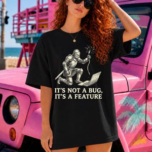 Puede incluir: Una camiseta negra de manga corta con un gráfico de un caballero con armadura arrodillado ante un portátil mientras sostiene un cable brillante. El texto impreso bajo el gráfico dice IT'S NOT A BUG, IT'S A FEATURE en letra clara y negrita.