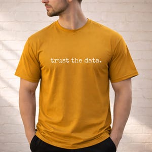 Puede incluir: Una camiseta de manga corta y cuello redondo de color amarillo mostaza con el texto blanco "trust the data." impreso en el pecho. La prenda está confeccionada en tela de color sólido y tiene un corte estándar con un diseño sencillo y limpio.