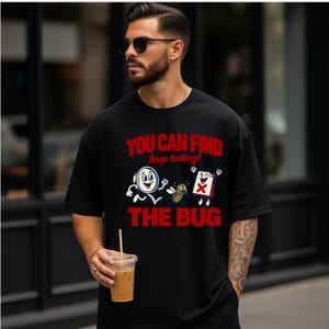 May include: A black short-sleeved t-shirt featuring a graphic print with the red text "YOU CAN FIND" and "THE BUG" surrounding a cartoon illustration of a magnifying glass, a small bug, and a document with a red X, plus the text "keep testing!"