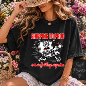 Peut inclure: Un t-shirt noir avec un motif de dessin animé représentant un ordinateur portable paniqué qui court en tenant une tasse de café renversée. Le texte SHIPPING TO PROD on a friday again est écrit en rouge et blanc avec un design ludique.