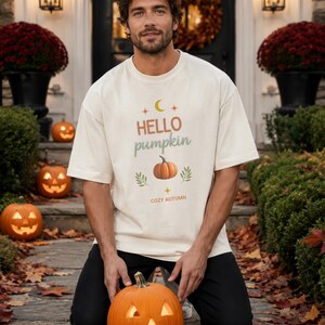 Puede incluir: Una camiseta de manga corta color hueso con un estampado gráfico que incluye una luna creciente amarilla, el texto "HELLO pumpkin" en naranja y verde, una ilustración de calabaza naranja, hojas verdes y el texto "COZY AUTUMN" en la parte inferior.