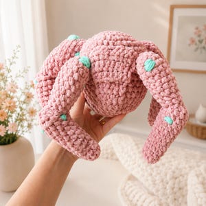 May include: A plush, crocheted spider toy crafted from soft, dusty pink yarn. The round body features eight thick, segmented legs extending outwards, each accented with small, bright turquoise yarn dots placed along the limbs and body.