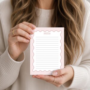 May include: A rectangular notepad featuring a light pink and white grid pattern background with a scalloped white border. The center contains horizontal black lines for writing and the name Millie printed in a light pink sans-serif font at the top.