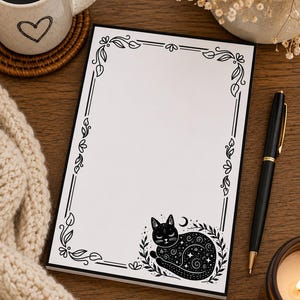 May include: A rectangular notepad with a white surface featuring a black printed border of leaves and vines. In the bottom right corner, there is a black illustration of a sleeping cat decorated with stars and swirls, surrounded by foliage.