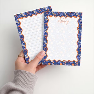 May include: Two rectangular stationery sheets featuring a dark blue border with a floral pattern of small white, orange, and light blue flowers. The border has a wavy orange edge. One sheet is lined, and the other has the name Ashley printed in orange.