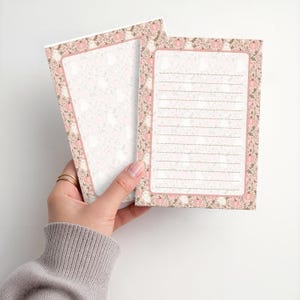 May include: Two rectangular stationery sheets featuring a border pattern of pink pumpkins, green foliage, and small white ghosts. One sheet is blank in the center, while the other contains horizontal lines for writing. The paper has a soft pink tone.