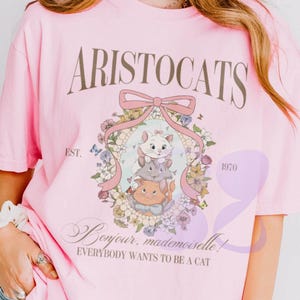 May include: A light pink short-sleeve t-shirt featuring a graphic of three kittens from The Aristocats inside a floral wreath with a pink bow. The text reads ARISTOCATS, EST. 1970, Bonjour, mademoiselle!, and EVERYBODY WANTS TO BE A CAT.