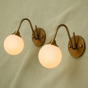 Könnte beinhalten: Ein Paar Wandleuchten mit runden, messingfarbenen Wandhalterungen und geschwungenen Armen. Jede Leuchte besitzt einen kugelförmigen Schirm aus mattem, weißem Glas, der ein weiches, diffuses Licht abgibt und für ein symmetrisches Erscheinungsbild sorgt.