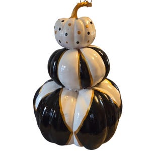 May include: A decorative stack of three pumpkins featuring a glossy finish. The top pumpkin is white with black and gold polka dots. The middle and bottom pumpkins feature alternating vertical black and white segments separated by thin gold lines.