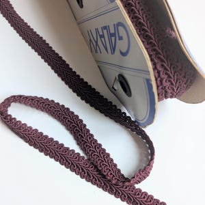 Peut inclure: Une bobine de garniture décorative de couleur mauve foncé, avec un motif bouclé détaillé. La garniture est partiellement déroulée, révélant le motif complexe. La bobine porte l'inscription "GALAXY".