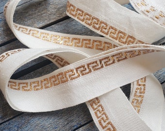 May include: White ribbon trim with a gold Greek key pattern. The ribbon is folded and lying on a blue wooden surface.