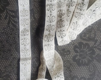 May include: White fabric trim with silver glitter accents. The trim features a repeating pattern of stylized floral designs.