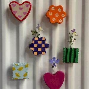 May include: A collection of six ceramic wall-mounted vases in various shapes and colors. The set includes heart, flower, and square forms featuring patterns like pink hearts, orange daisies, purple and peach checkers, green stripes, and yellow lemons.
