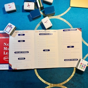 May include: A 2026 Mah Jongg Line Tracker score card with sections for consecutive runs, winds, dragons, and numbers, surrounded by scattered white and blue Mah Jongg tiles on a blue surface with a gold-colored corner piece visible.