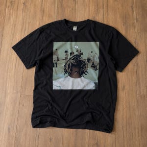 Puede incluir: Una camiseta negra de manga corta con un estampado gráfico central. La imagen muestra a una persona con rastas rubias vista desde atrás, con una camisa blanca, en una sala clínica con varias figuras con batas blancas.