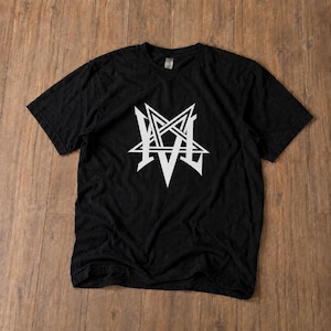 Puede incluir: Una camiseta negra de manga corta con un gran estampado gráfico blanco en el pecho. El diseño consiste en una estrella de cinco puntas estilizada, o pentagrama, integrada con las letras M y V, creando un emblema geométrico y simétrico.