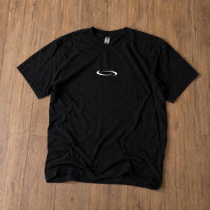 Puede incluir: Una camiseta negra de manga corta con un pequeño logotipo ovalado estilizado en color blanco centrado en el pecho. La prenda tiene un cuello redondo clásico y un corte estándar, confeccionada en un tejido negro liso con etiqueta visible.