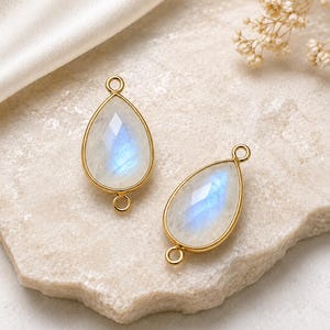 Peut inclure: Deux breloques en pierre de lune en forme de goutte serties dans des cadres en métal doré. Chaque breloque présente une surface facettée avec un éclat irisé bleu et comprend deux petites boucles circulaires en haut et en bas pour la fixation.