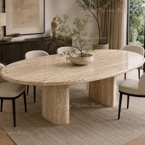 May include: An oval dining table featuring a polished beige travertine stone top and two matching cylindrical travertine pedestal bases, surrounded by four off-white upholstered chairs with dark wooden legs and a small plant in a bowl.