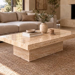 Pode incluir: Uma mesa de centro retangular de travertino bege com superfície polida e base de bloco sólido. A mesa apresenta uma textura de pedra porosa natural e assenta sobre um tapete castanho texturizado. A superfície contém dois livros, uma taça e um vaso.