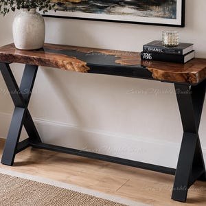 May include: A console table featuring a live-edge wood slab with a central black resin inlay, supported by a black metal X-frame base. The tabletop displays natural wood grain patterns and textures, contrasting with the smooth, dark resin surface.