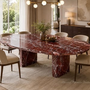 May include: An oval dining table featuring a deep red marble top with white veining, supported by two matching red marble pedestal bases. The table is surrounded by six beige upholstered chairs with dark wooden legs, arranged in a dining room.