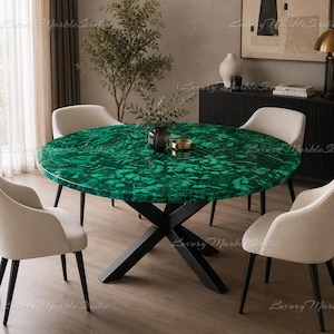 Può includere: Un tavolo da pranzo rotondo con un piano dal vivace motivo in malachite verde e finitura lucida, sostenuto da una base in metallo nero a forma di X. Il tavolo è circondato da quattro sedie imbottite color bianco sporco con gambe nere affusolate.