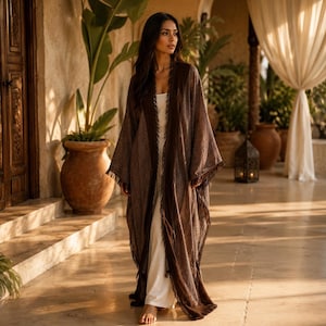 Peut inclure: Un long kimono marron ouvert sur le devant, doté d'un tissu texturé avec des bords effilochés le long des revers et de l'ourlet, porté sur une robe blanche simple arrivant au sol. Le vêtement a des manches larges et une coupe fluide.