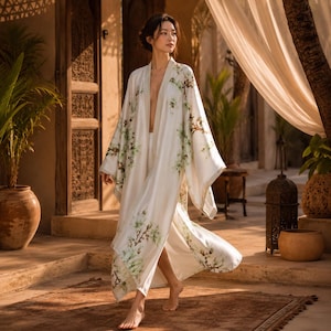 May include: A long, flowing, cream-colored robe featuring an open front and wide sleeves. The fabric is decorated with a delicate, repeating pattern of green leaves and brown branches, creating a soft, botanical aesthetic across the entire garment.