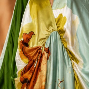 May include: A satin dress featuring a vibrant abstract print in shades of olive green, light blue, white, and burnt orange. The garment has a gathered V-neckline with soft, fluid folds and a smooth, lustrous finish that drapes across the fabric.