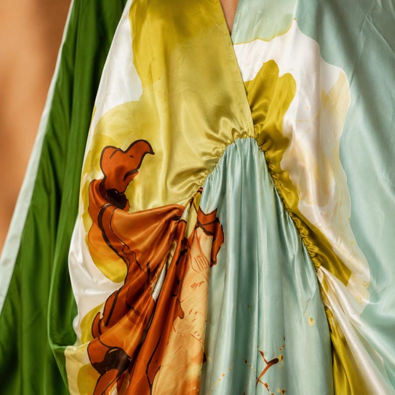 May include: A satin dress featuring a vibrant abstract print in shades of olive green, light blue, white, and burnt orange. The garment has a gathered V-neckline with soft, fluid folds and a smooth, lustrous finish that drapes across the fabric.
