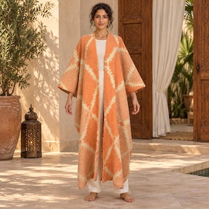 Puede incluir: Un cárdigan largo de textura abierta en color naranja y crema con un patrón de diamantes, usado sobre una blusa blanca sencilla y pantalones blancos. La prenda tiene mangas anchas de tres cuartos y una silueta fluida.