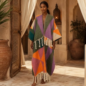 May include: A long, open-front kimono-style garment featuring a geometric pattern of large triangles in shades of olive green, vibrant orange, purple, and cream. The piece is finished with thick, cream-colored twisted fringe along the hemline.