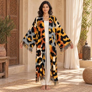 May include: A long, open-front kimono-style cardigan featuring a bold leopard print in shades of mustard yellow, black, and grey. The garment has wide, three-quarter length sleeves and a straight hemline finished with long, decorative fringe trim.