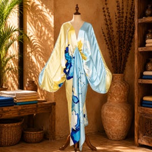 May include: A floor-length satin dress featuring a deep V-neckline and wide, flowing sleeves. The garment displays a vibrant abstract pattern in shades of pale yellow, sky blue, and deep royal blue, with a gathered waist detail.
