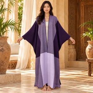 May include: A long, open-front kimono-style cardigan featuring a colour-blocked design in shades of deep purple, medium violet, and light lavender. The garment has wide, flowing sleeves and a textured knit pattern throughout the fabric.