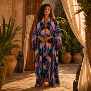 May include: A long, open-front crochet cardigan featuring a vibrant geometric pattern in shades of blue, pink, yellow, and dark purple. The garment has long sleeves, a shawl collar, and a hemline finished with long, matching multicolored fringe.