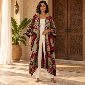 May include: A long, open-front hooded cardigan featuring a beige base with a bold geometric pattern in red, black, and grey. The garment has wide sleeves and a straight hem, worn over a plain white tank top and matching white trousers.