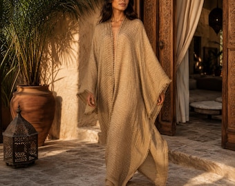 May include: A floor-length, open-front kaftan in a textured, light tan fabric. The garment features wide, loose sleeves, a straight silhouette, and a delicate, frayed edge trim along the front opening and hemline.