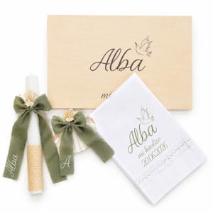 Puede incluir: Una vela blanca con lazo verde salvia y flores secas, una concha con lazo verde salvia y flores secas, y un paño blanco con encaje, todo con el nombre Alba y una paloma, junto a una caja de madera con el nombre Alba impreso en la superficie.