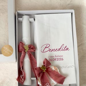 Puede incluir: Una vela de bautizo blanca y una servilleta de tela blanca dentro de una caja blanca. La vela tiene un borde de encaje blanco, un lazo rosa empolvado y un dije de paloma dorada. La servilleta lleva bordado el nombre Benedita, o meu batismo, 26.09.2026.