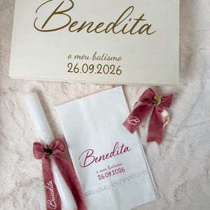 Puede incluir: Una caja de recuerdos de madera blanca, una vela blanca y un paño blanco, todos personalizados con el nombre Benedita, la frase o meu batismo y la fecha 26.09.2026. La vela y el paño incluyen cintas de terciopelo rosa empolvado y encaje.