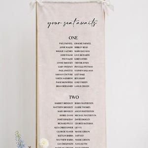 Puede incluir: Un cartel de distribución de asientos rectangular y alto de tela beige, suspendido de un marco de metal dorado con lazos blancos. El cartel presenta el texto 'your seat awaits' en letra negra, seguido de nombres bajo los títulos ONE y TWO.
