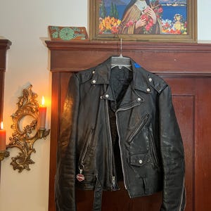 May include: Black leather motorcycle jacket with silver zippers, snaps, and a belt. The jacket has a classic asymmetrical design and is hanging on a hanger. A Harley Davidson keychain hangs from the zipper. A painting and a sign are in the background.