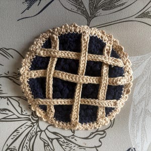May include: A circular, crocheted coaster designed to resemble a pie. The centre features a dark navy blue textured yarn representing the filling, while the top is adorned with a beige lattice crust pattern and a scalloped, crocheted border.