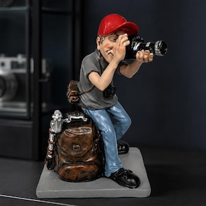 Puede incluir: Una figura de un fotógrafo con gorra roja, camiseta gris y vaqueros azules, sosteniendo una cámara negra frente al ojo. La figura está sentada sobre una mochila marrón con un trípode plateado al lado, sobre una base rectangular gris.