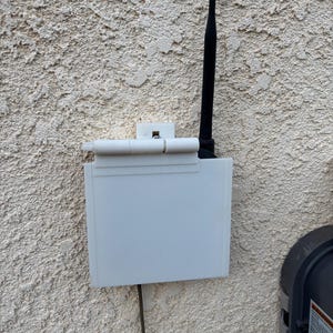 May include: A white rectangular plastic protective cover mounted on a textured wall, featuring a hinged top flap and a black vertical antenna extending from the right side, with a thin dark cable extending downward from the bottom of the unit.
