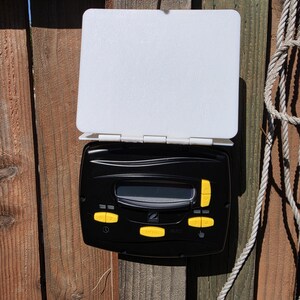 May include: A black electronic control unit with a digital display screen and yellow buttons, featuring a white hinged protective cover mounted above the main interface. The device includes directional arrows and menu selection buttons.