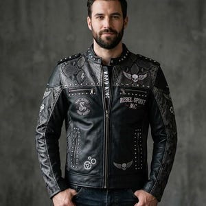 May include: A black leather jacket featuring silver studs, grey snakeskin-patterned panels, and various patches including a skull with wings, a cobra emblem, and text reading "REBEL SPIRIT MC" and "ROAD KING" along the front zipper closure.