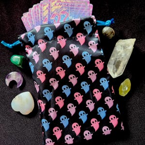 May include: A black drawstring pouch featuring a repeating pattern of small pink and light blue ghosts. The pouch contains a deck of iridescent cards and is surrounded by various polished crystals, including a clear quartz point and a moon-shaped stone.